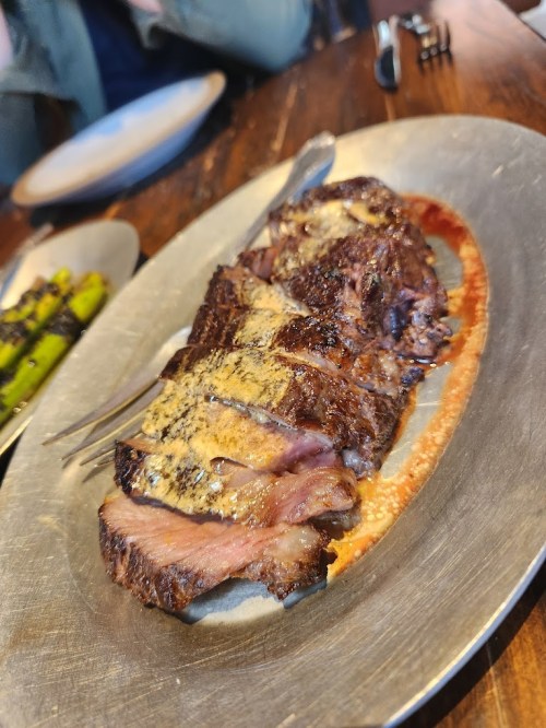 A platter of sliced, grilled steak with a golden crust, served with a side of asparagus on a wooden table.