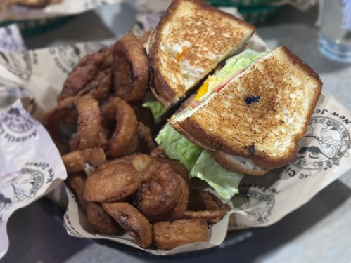 A toasted sandwich with lettuce and tomato, served with a side of crispy onion rings in a basket.