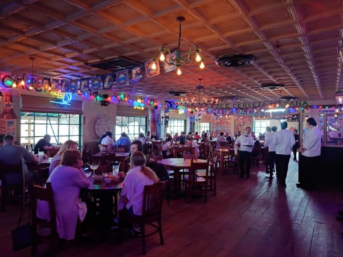 A lively restaurant interior with colorful lights, diners at tables, and staff serving in a warm, inviting atmosphere.