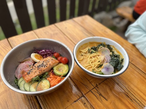 Two bowls of colorful dishes on a wooden table: one with salmon, vegetables, and an egg; the other with noodles in a creamy sauce.