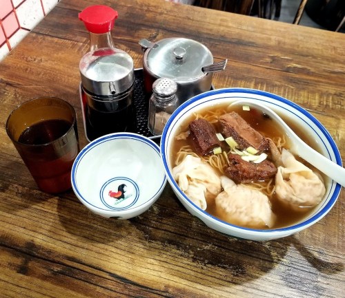 A bowl of noodle soup with dumplings and meat, accompanied by condiments and a drink on a wooden table.
