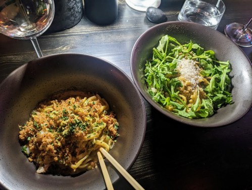 Two bowls of food: one with pasta topped with breadcrumbs and herbs, the other with arugula and grated cheese.