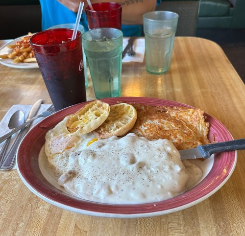 A plate with biscuits and gravy, eggs, and a toasted bun, with drinks in the background on a wooden table.