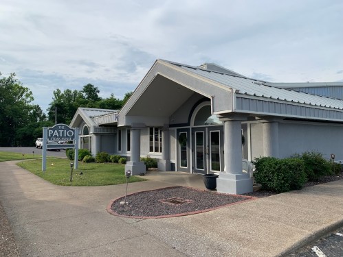 Exterior view of a building with a sloped roof, featuring large windows and a sign for "The Patio." Green landscaping surrounds it.