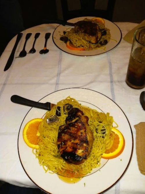 Two plates of grilled chicken on a bed of spaghetti, garnished with orange slices, on a table with utensils.