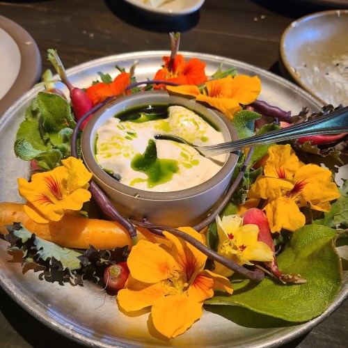A vibrant platter of colorful flowers, greens, and a creamy dip drizzled with green oil, garnished with radishes and carrots.