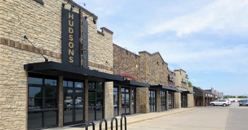 A row of stone storefronts with large windows, featuring the sign "HUDSON'S" and a clear blue sky above.