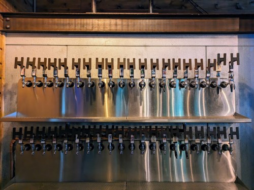 A wall-mounted beer tap system with multiple taps arranged in two rows, reflecting light from a nearby source.