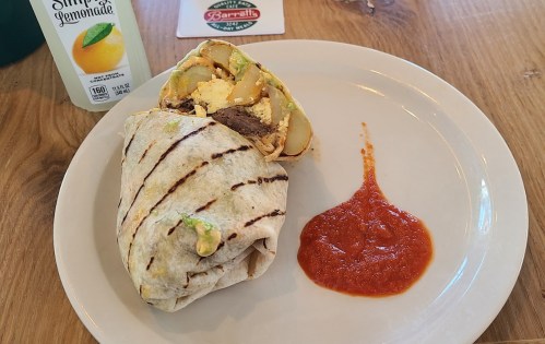 A grilled burrito filled with meat and vegetables, served with a side of red sauce and a drink of lemonade.