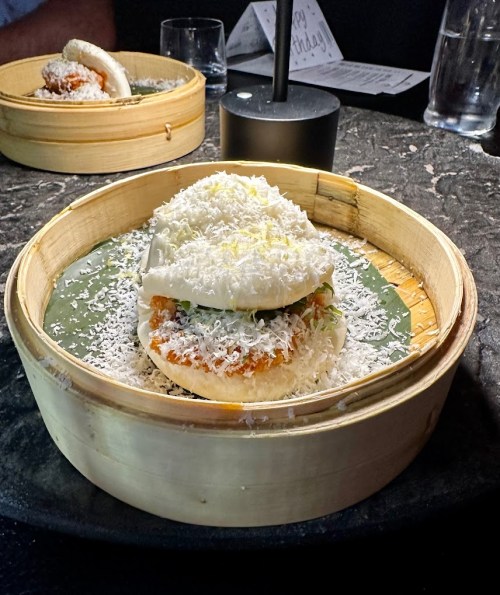 A bamboo steamer holds a bao bun filled with crispy chicken, topped with shredded cheese and garnished with herbs.