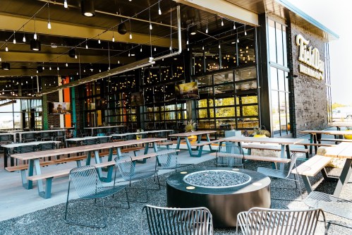 Outdoor dining area with wooden tables, chairs, and a fire pit, featuring a modern building with large windows.
