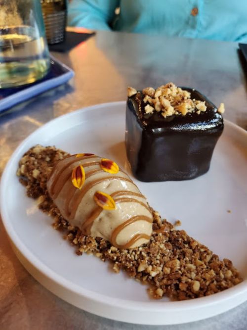 A dessert plate featuring a creamy mousse with drizzled sauce and a chocolate cube topped with crushed nuts.