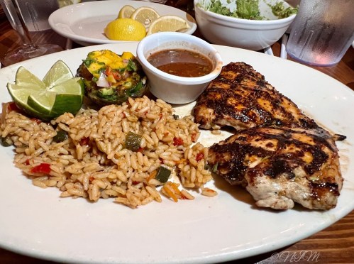 Grilled chicken with rice, a stuffed pepper, dipping sauce, and lime wedges on a white plate.