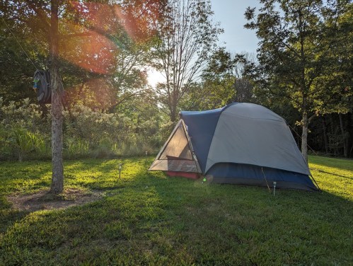 A tent set up on green grass, surrounded by trees, with sunlight filtering through the leaves in the background.