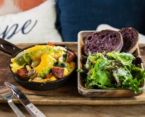 A skillet dish with vegetables and cheese, served with a side salad and slices of purple bread on a wooden tray.