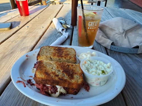 A plate with a toasted sandwich, coleslaw, and a glass of iced tea on a wooden table outdoors.