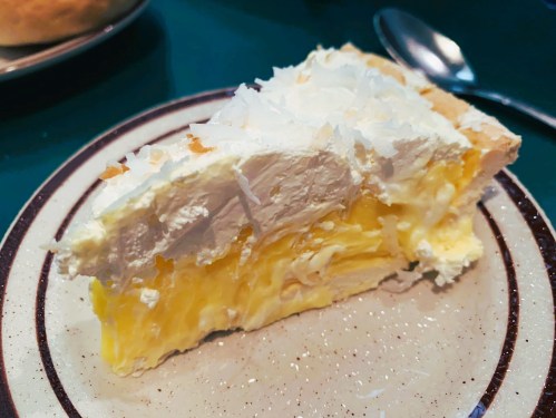 A slice of creamy lemon pie topped with whipped cream on a speckled plate, with a spoon nearby.