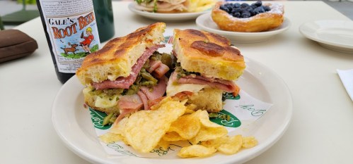 A sandwich with ham and pickles, served with chips and a bottle of root beer, with a pastry in the background.