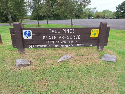 Sign for Tall Pines State Preserve in New Jersey, featuring environmental protection symbols and a grassy area.