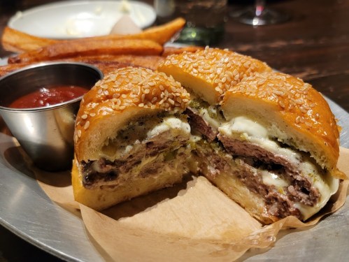 A close-up of a sesame seed burger bun filled with a juicy beef patty, melted cheese, and served with fries and ketchup.