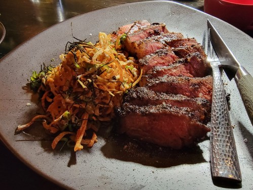 A plate featuring sliced grilled steak alongside a serving of crispy fried noodles and garnished vegetables.