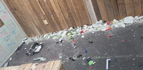 A messy area with broken glass and debris scattered on the ground near a wooden fence.