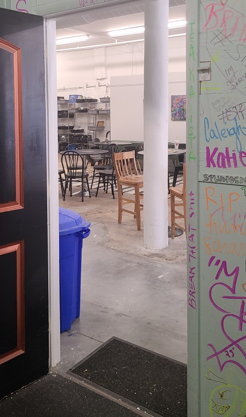 View through an open door into a spacious room with tables, chairs, and a blue trash bin, featuring colorful graffiti on the walls.