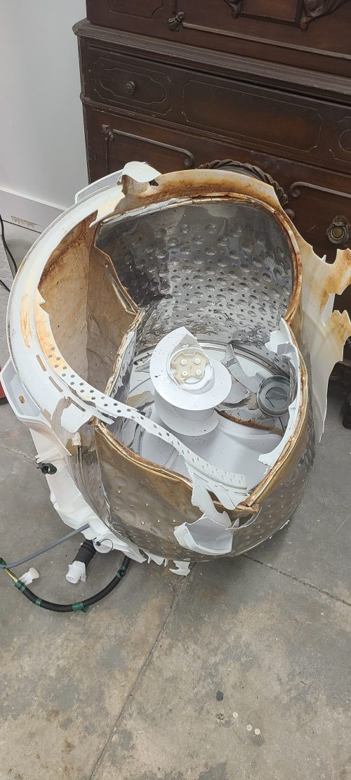A disassembled washing machine drum showing rust and damage, with internal components exposed on a concrete floor.