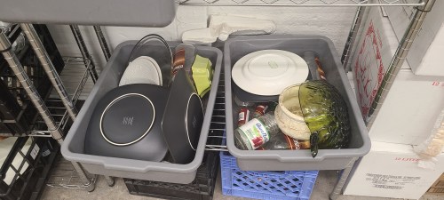 Two gray bins filled with plates, bowls, and canned goods, organized on a metal shelf in a storage area.
