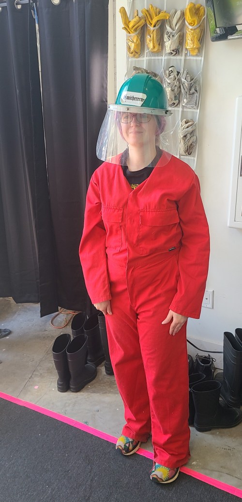 A person in a red jumpsuit and safety helmet stands in a room, wearing a face shield and colorful footwear.