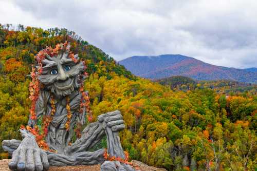 A whimsical tree-like sculpture with a face, surrounded by vibrant autumn foliage and mountains in the background.