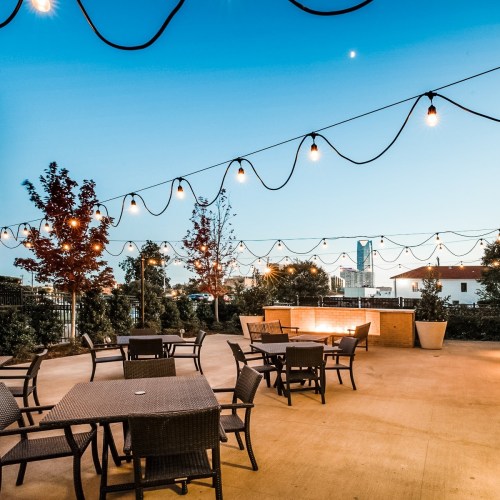 Outdoor seating area with string lights, trees, and a city skyline in the background during twilight.
