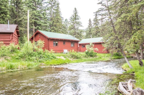 Two red cabins beside a clear stream, surrounded by lush greenery and tall trees in a serene natural setting.