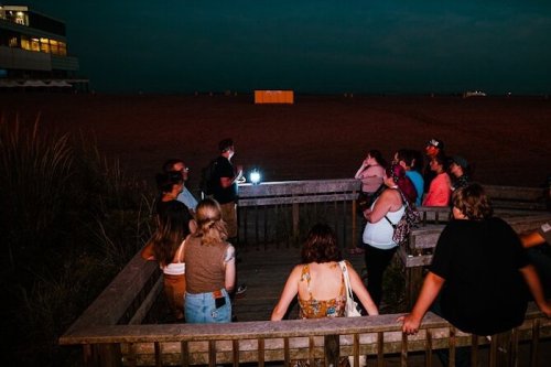 A group of people gathered on a deck at dusk, listening to a speaker with a light, overlooking a beach.