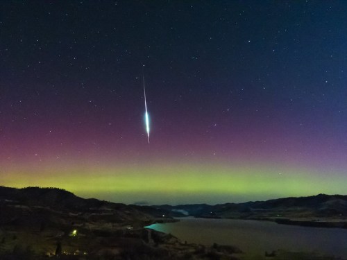 A stunning night sky with a bright meteor streaking above a colorful aurora over a calm lake and hills.