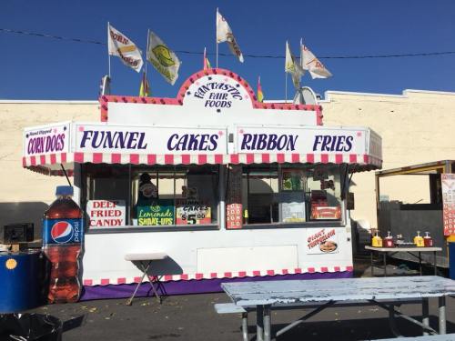 A colorful fair food stand with flags, offering funnel cakes, ribbon fries, and corn dogs, with picnic tables nearby.