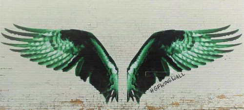 A vibrant mural of green wings spread wide on a white brick wall, featuring the hashtag #GFwingwall.