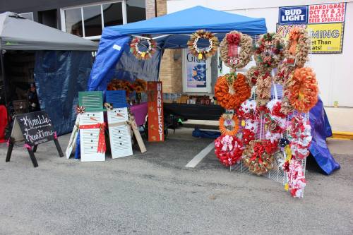 A colorful market stall with wreaths and decorations under a blue canopy, featuring a sign that says "Harvest Greetings."