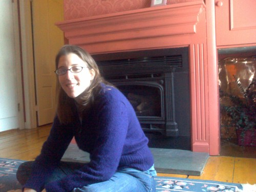 A woman with glasses sits on the floor in a cozy room with a fireplace and pink walls.