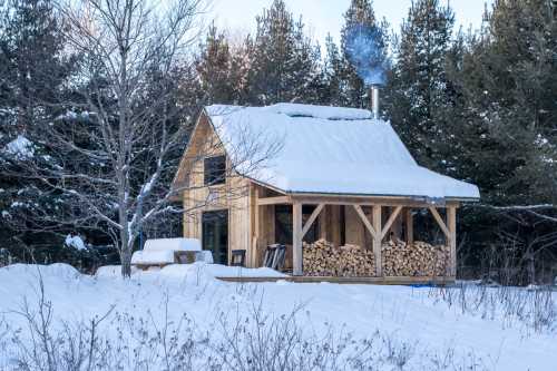 A cozy wooden cabin covered in snow, with a chimney emitting smoke and stacked firewood in front. Pine trees in the background.