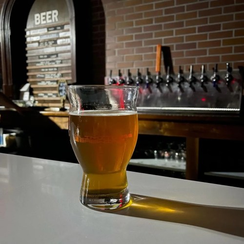 A glass of beer sits on a bar counter, with a brick wall and beer taps in the background.