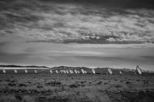 A row of satellite dishes in a grassy field, set against a cloudy sky and distant mountains, in black and white.