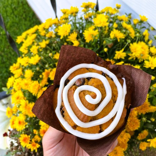 A hand holding a pumpkin muffin with white icing swirls, set against a backdrop of bright yellow flowers.