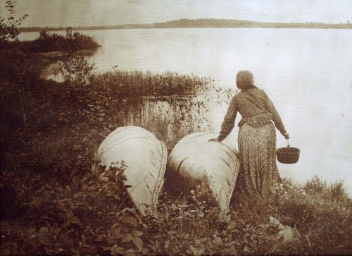 A sepia-toned image of a woman in a long dress by a river, standing beside two canoes and holding a basket.