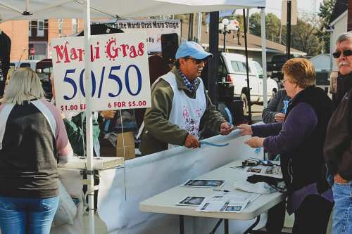 A vendor at a booth sells 50/50 raffle tickets during a festival, with a sign displaying prices and event details.