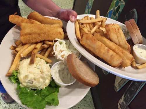 Two plates of fried fish with French fries, coleslaw, potato salad, and a slice of bread.