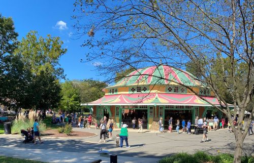 A colorful carousel building surrounded by trees and people enjoying a sunny day at a park.