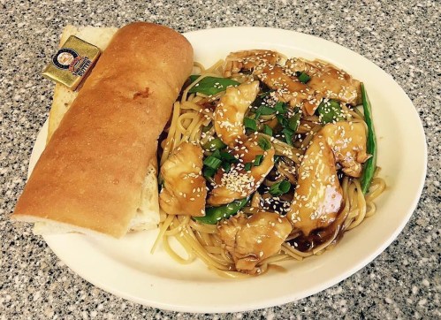 A plate of chicken stir-fry with noodles, topped with sesame seeds, alongside a piece of bread and butter.