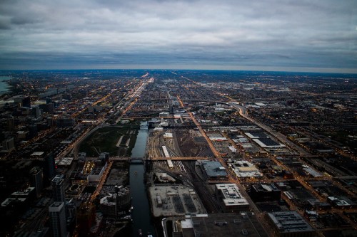 Aerial view of a cityscape at dusk, featuring roads, buildings, and a river under a cloudy sky.