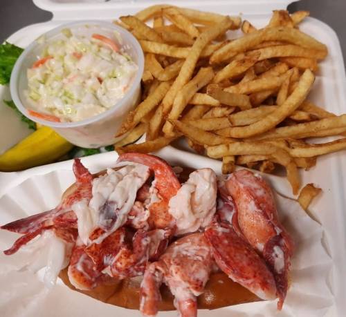 A takeout container with lobster meat, fries, coleslaw, and a pickle on the side.